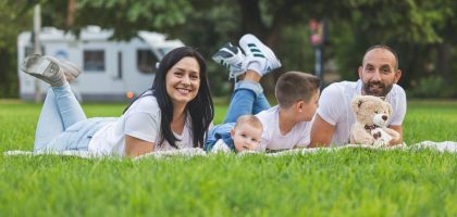 Happy family lying on grass with RV in the background, enjoying a sunny outdoor picnic.